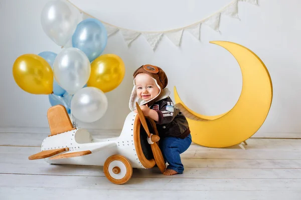 A happy one-year-old boy in a helmet and a pilot\'s jacket stands near a wooden plane, next to balloons and a yellow moon. Portrait in the studio of a child aviator.