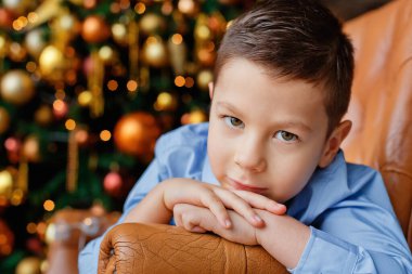 Mavi gömlekli bir çocuk, arka planda altın bir Noel ağacının ve çelenk ışıklarının önünde koltukta oturmuş, rüyasında kameraya bakıyor. Seçici odak