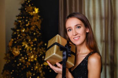 Altın dekoru olan bir Noel ağacının arka planında siyah elbiseli zarif bir bayan. Genç bir kadın elinde siyah kurdeleli altın bir paketle bir hediye tutuyor..