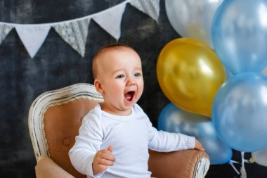 Stüdyoda küçük bir çocuğun portresi. Çocuk, bayraklardan oluşan bir çelenkle süslenmiş koyu gri arka planda bir grup balonun yanında mini sandalyede oturuyor..