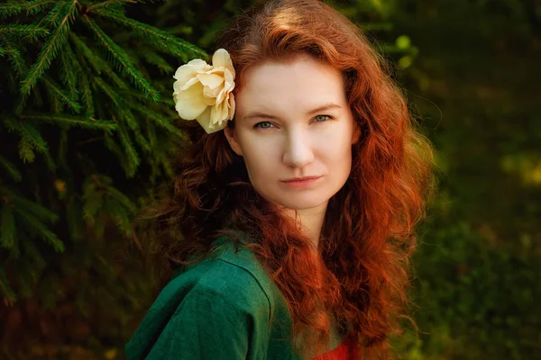 stock image Portrait of a red-haired woman with a rose flower in her hair in the park. A curly-haired woman near the green fir branches.