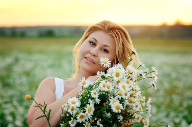A large portrait of a young, plump, beautiful woman resting on a chamomile field at sunset. A plus-size woman in a white sundress hugs a large bouquet of flowers in a meadow with daisies.