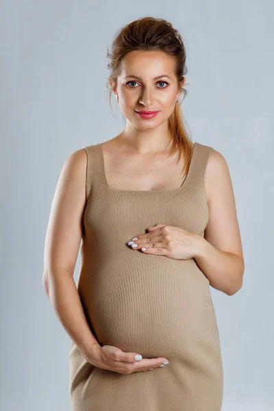 一个穿着米黄色衣服的孕妇的特写肖像 双手抱在灰色背景上 温柔地看着她的胃 等着分娩 — 图库照片