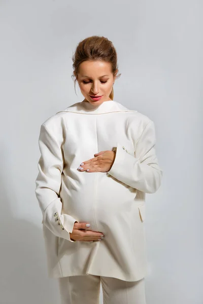 穿着米色西服 背着夹克 站在灰色工作室背景上 穿着怀孕时尚的漂亮的怀孕金发女商人妈妈 — 图库照片