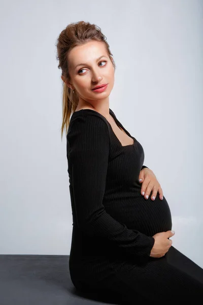 工作室里一个穿着黑色衣服 身披灰色背景的金发孕妇的时髦画像 她坐着 双手轻轻地握住她的胃 — 图库照片