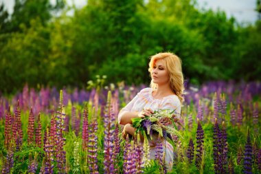 Genç, tombul bir sarışının portresi. Bir kadın gün batımında çayırda bir demet leylak pembesi çiçek topluyor..
