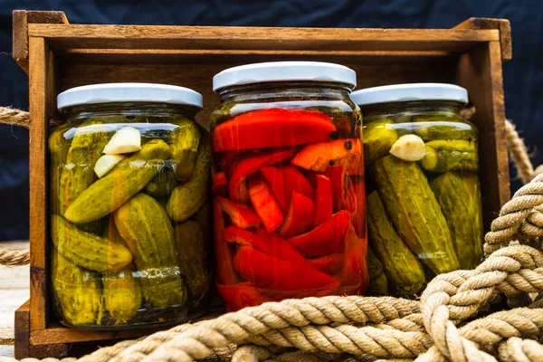 Glass jars with pickled red bell peppers and pickled cucumbers (pickles) isolated in wooden crate. Jars with variety of pickled vegetables. Preserved food concept in a rustic composition.