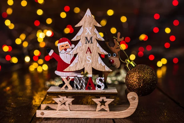 Arka planda bulanıklık ve odak ışıkları karşısında Noel süslemeleri. Noel Baba, ren geyiği, Noel ağacı ve ışıltılı Noel toplarıyla ahşap bir Noel kızağı çek.