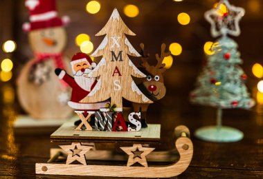 Arka planda bulanık bir Noel süsü var. Noel Baba, ren geyiği ve Noel ağacı olan ahşap bir Noel kızağının önünde..