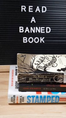 Reminder to read a banned book, along with copies of oft-banned books To Kill a Mockingbird, Stamped, and Of Mice and Men.