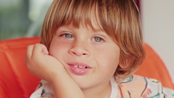 Primer plano del niño de ojos azules, sentado en una silla de color naranja, en una mañana soleada — Vídeos de Stock