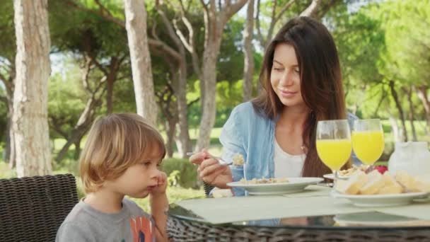 Netter Junge genießt sein Frühstück und fruchtigen Drink, während er neben seiner Mutter sitzt — Stockvideo