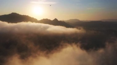 Altın gün batımında bulutların ve dağların üzerinde uçan küçük turistik helikopter 4K