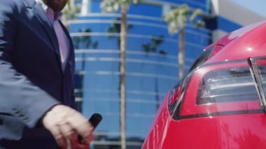 İş adamı güneşli yaz gününde elektrikli arabayı şarj ediyor. Kapağın altından alçak açılı çekim.