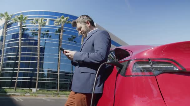 Man in blauw pak, staande bij het opladen van elektrische auto, werken op smartphone — Stockvideo