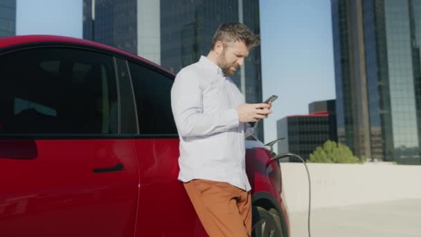 Amerikansk affärsman använder smartphone och lutar sig mot att ladda lyx elbil — Stockvideo