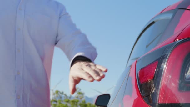 El hombre conecta un coche eléctrico al cargador en los generadores de viento en el fondo de movimiento — Vídeos de Stock