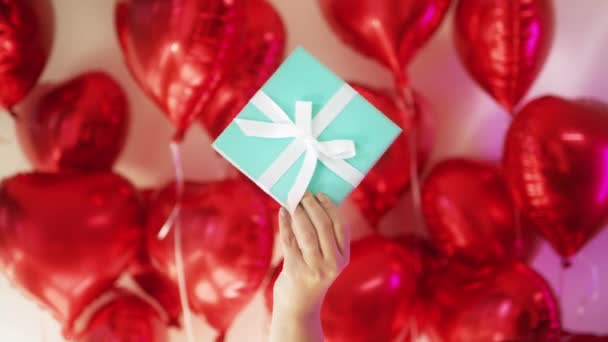 Concepto de amor, mano femenina con caja azul cinta blanca en el fondo de globos rojos — Vídeo de stock