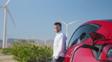 Elektrikli araba şarjının fişini çeken adam birkaç rüzgar jeneratörü döndürüyor.
