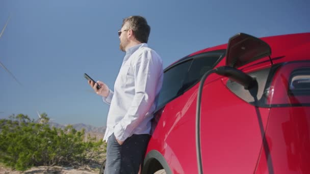 起業家はスマートフォンをチェックし電気自動車の充電を待っています — ストック動画