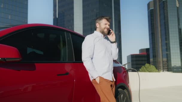 Бізнесмен використовує смартфон під час зарядки електромобіля на офісному паркінгу — стокове відео