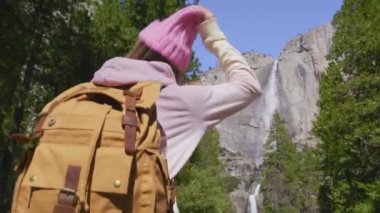 Mutlu, gülümseyen bir kadın sırt çantasıyla Yosemite şelalesine yürüyor.