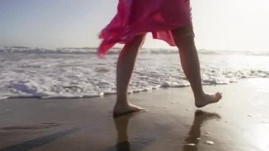 Kırmızı kamera beyaz dalgalarla deniz kıyısında yürüyen kadının bacaklarını çekiyor.