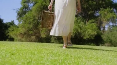 Piknik sepeti ile şehir parkında yürüyen beyaz boho elbiseli kadın.