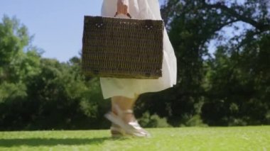 Genç bir kadın, elinde sepet, yanında da kırmızı kamerayla orman bahçesinde yürüyor.