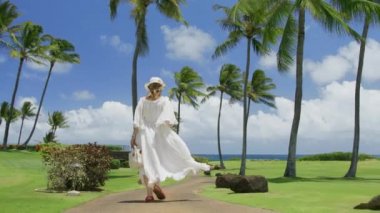 Rüya gezisi, tatil gezisi konsepti. Mükemmel bir yaz tatili gününde lüks bir tatil köyünde yürüyen mutlu özgür bir kadın. Beyaz sallanan elbise ve şapkalı kadın tropikal adada, RED 6K çekti.