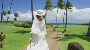 Rüzgarda pohpohlayan muhteşem beyaz elbiseli genç kadının yavaş çekim arkası görüntüsü, tropik Hawaii adasında lüks bir tatil köyünün önünden geçen kadın. Sinematik yaz manzarası, tatil seyahati konsepti kırmızı