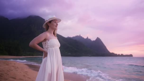 考艾岛4K红顶绿油油的Napali海岸公园山顶上 穿着雅致的白帽 穿着华丽的漂亮衣服 带着紫色云彩 享受着电影般的粉色夕阳的美丽女性形象 — 图库视频影像