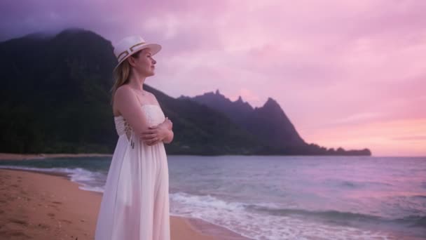 エレガントな白いドレスで豪華な笑顔の美しい女性の肖像画は壮大な緑と紫の海の上の映画のピンクの黄金の夕日を楽しんでいますナパリ海岸の山のピークカウアイ島ハワイ島4K Red — ストック動画