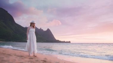 Güzel, gülümseyen bir kadın, Kauai Adası 'ndaki NaPali sahillerinde, pembe mor bulutların üzerinde, gün batımında, çıplak ayakla sinematik kumlu plajda yürüyor. Hawaii 4K 'de destansı yaz tatili.
