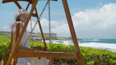 Okyanus manzaralı lüks sahil tatil köyü parkında ahşap salıncakta sallanan genç bir kadın. Mutlu yaz tatili hayalleri. Tahta salıncakta zarif bir bayan Hawaii 'de tatilin tadını çıkarıyor.
