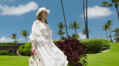 Yavaş çekim gülümseyen kadın güneşli yaz gününde tropik adanın yanından geçiyor. Uzun beyaz dalgalı elbise, deniz esintisinde uçuşan sarı saçlar. Mutlu turistler Hawaii 'de tatilin tadını çıkarıyor.
