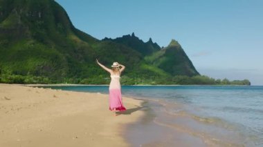 Gün doğumunda okyanus sahilinden dağ tepelerine doğru koşan renkli elbiseli, kollarını havaya kaldıran mutlu ve hayat, gençlik ve mutluluktan sarhoş bir kadın. Hawaii 'nin tadını çıkarıyorum. Güzel manzara.