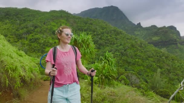 年轻微笑的女人带着背包和手杖在夏日的热带雨林中穿行 考艾岛 在多云炎热的热带天气 沿着布满绿色丛林的电影般的山地海岸徒步旅行 — 图库视频影像