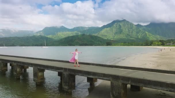 スローモーション若い女性の動きの背景に緑の山のピークとビーチ桟橋で歩くドレスを放棄し 空気中の腕を上げ 幸せな生活夢の生活 若者と幸福 ハワイの自然を楽しむ — ストック動画