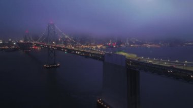 Çok katmanlı modern asma köprüyle dönen arabalar karanlık gece, San Francisco 'da aydınlandı. Sinematik endüstriyel çekim. Yoğun şehir yolunda gece vakti 4K hava sahası çöktü.