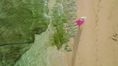 Drone, canlı pembe elbiseli ve zarif şapkalı gezgin kadını takip ediyor. Hawaii adasındaki okyanus kıyısındaki tropikal sahilde yürüyen ağır çekimde bir kadın. Güneş ışınları dalgalanan su yüzeyinde parlıyor.