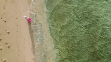 Hawaii adasındaki okyanus kıyısındaki tropikal sahilde yürüyen ağır çekimde bir kadın. Güneş ışınları parıldayan su yüzeyini aydınlatıyor. Drone pembe elbiseli ve zarif şapkalı gezgin kadını takip ediyor.