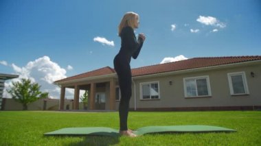 Orta yaşlı fitness kadını yeşil çimlerin üzerinde egzersiz yapıyor. Avrupalı bir kadın güneşli bir günde evinin arka bahçesinde çömeliyor. O gülümsüyor. Yüksek kalite 4k görüntü