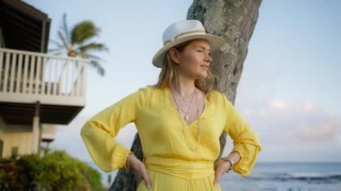 Gün batımında okyanus manzaralı, parlak sarı elbiseli, ağır çekim gülümseyen kadın. Özgürlük hissi, yaz tatili, rüya yolculuğu, sahil doğası kavramı. Kırmızı Hawaii adasına 6K yolculuk