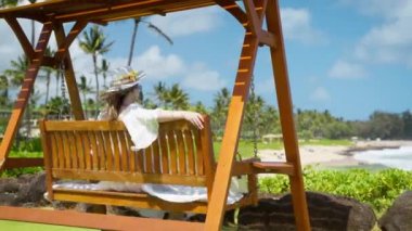 Tahta salıncaktaki sarışın kadın Hawaii Adası 6K 'da tatilin tadını çıkarıyor. Mutlu yaz tatili hayalleri. Şık bir bayanın ahşap salıncakta sallanışının arkası lüks okyanus manzaralı bir tatil köyünde