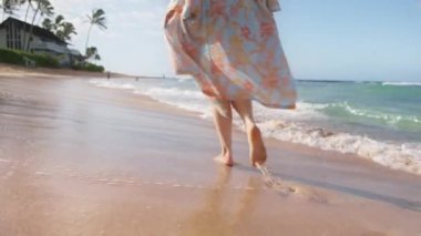Yavaş hareket eden kadın ayaklarıyla sahilde yalın ayak yürüyor altın gün doğumunda okyanus dalgalarıyla yıkanmış ıslak kumda ayak izleri bırakıyor. Hawaii adasında yaz tatilindeki kadın turist. Yazlık plaj elbiseli kadın.