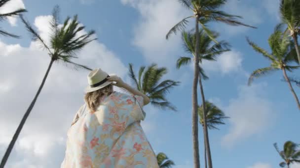 低角美女模型在度假 穿着五彩缤纷的海滩和服的慢动作的年轻女子仰望着热带岛屿棕榈树和蓝天 飞行夏装中迷人的时尚模特 — 图库视频影像