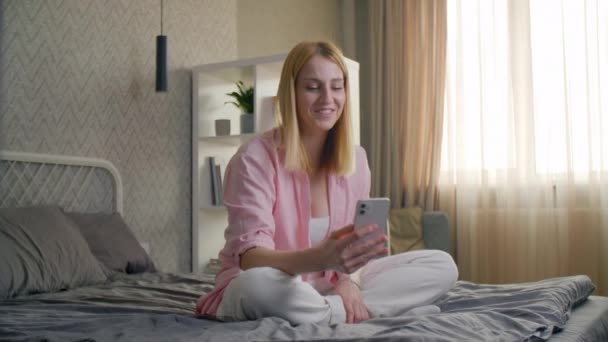 女人用智能手机和朋友进行远程交谈 白人金发姑娘坐在舒适时髦的房间里的床上 高质量的4K镜头 — 图库视频影像