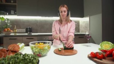 Bir kadının müzik dinlerken salatalık dilimlemesi. Beyaz kız mutfağında salata yapıyor. Yüksek kalite 4k görüntü