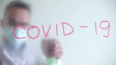 İçerideki tecrit döneminde yetişkin bir adam. Adam cam bir tahtadan Covid-19 sembolünü çıkarmak için sıvı püskürtür. Yüksek kalite 4k görüntü